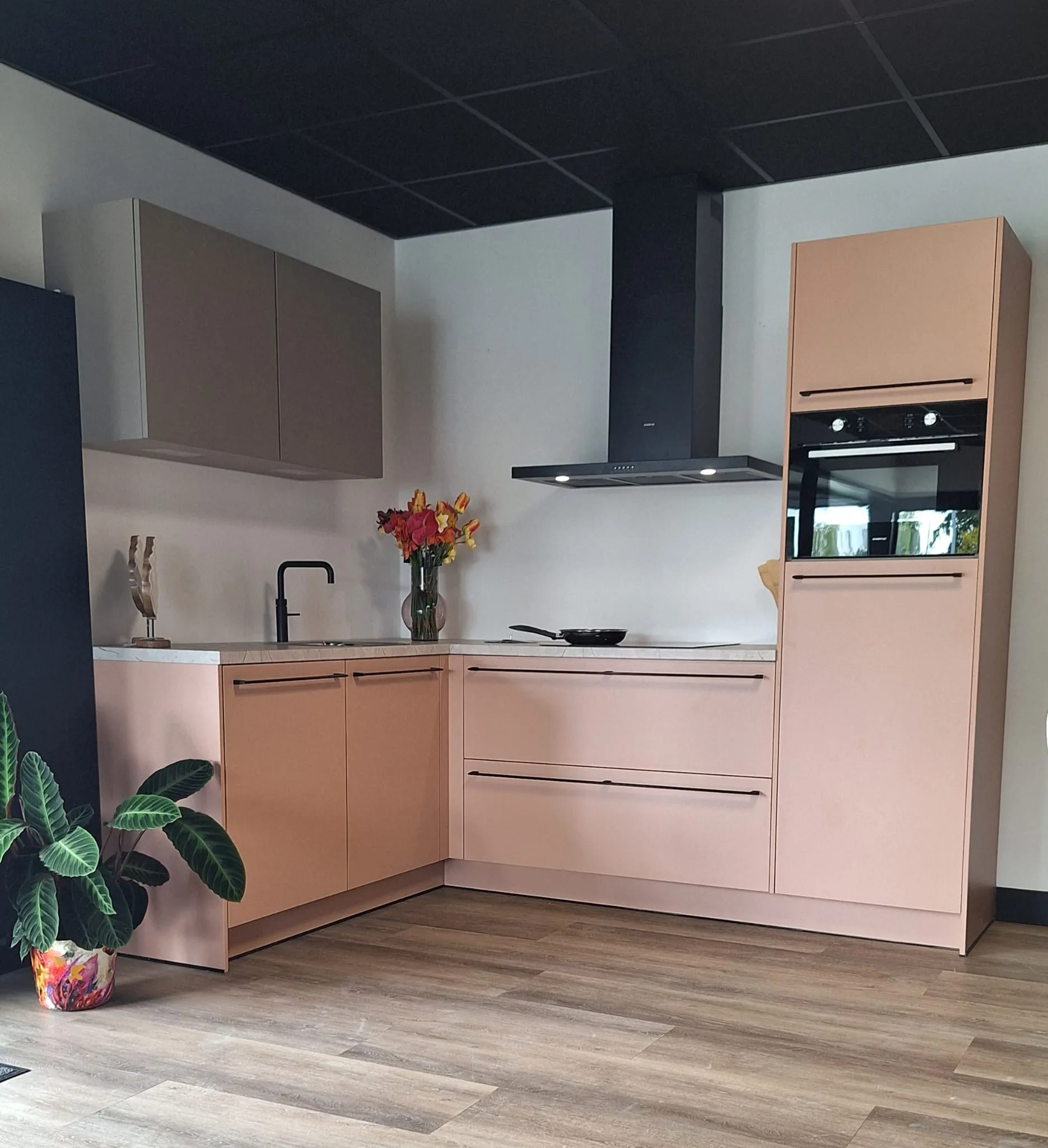 Hoekkeuken in zachtroze met taupe bovenkasten, matzwarte afzuigkap en inbouwoven.